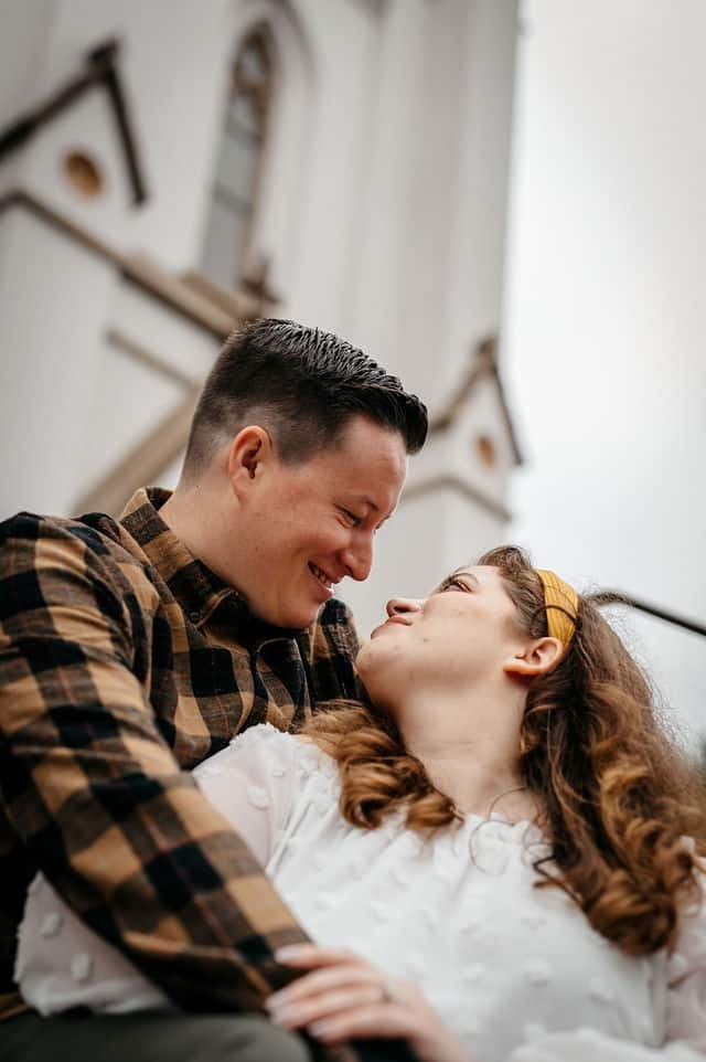 An engaged couple laughing together on the steps of a white church.