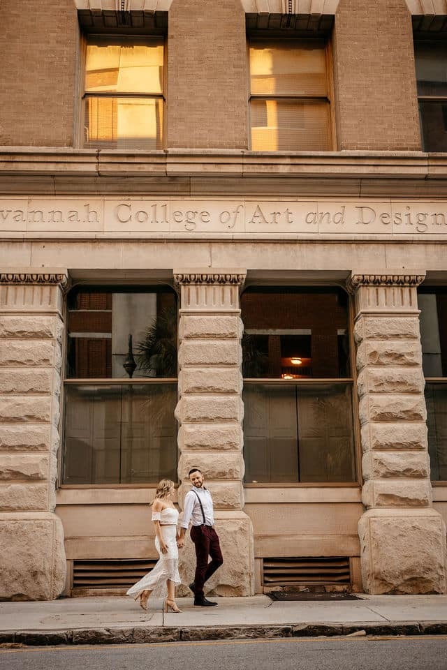 An engaged couple walking hand in hand past a historic Savannah facade.