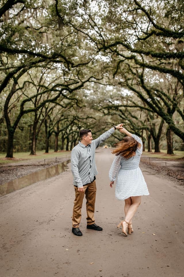 A man twirling his fiancee on a dirt lane beneath moss-draped oaks.