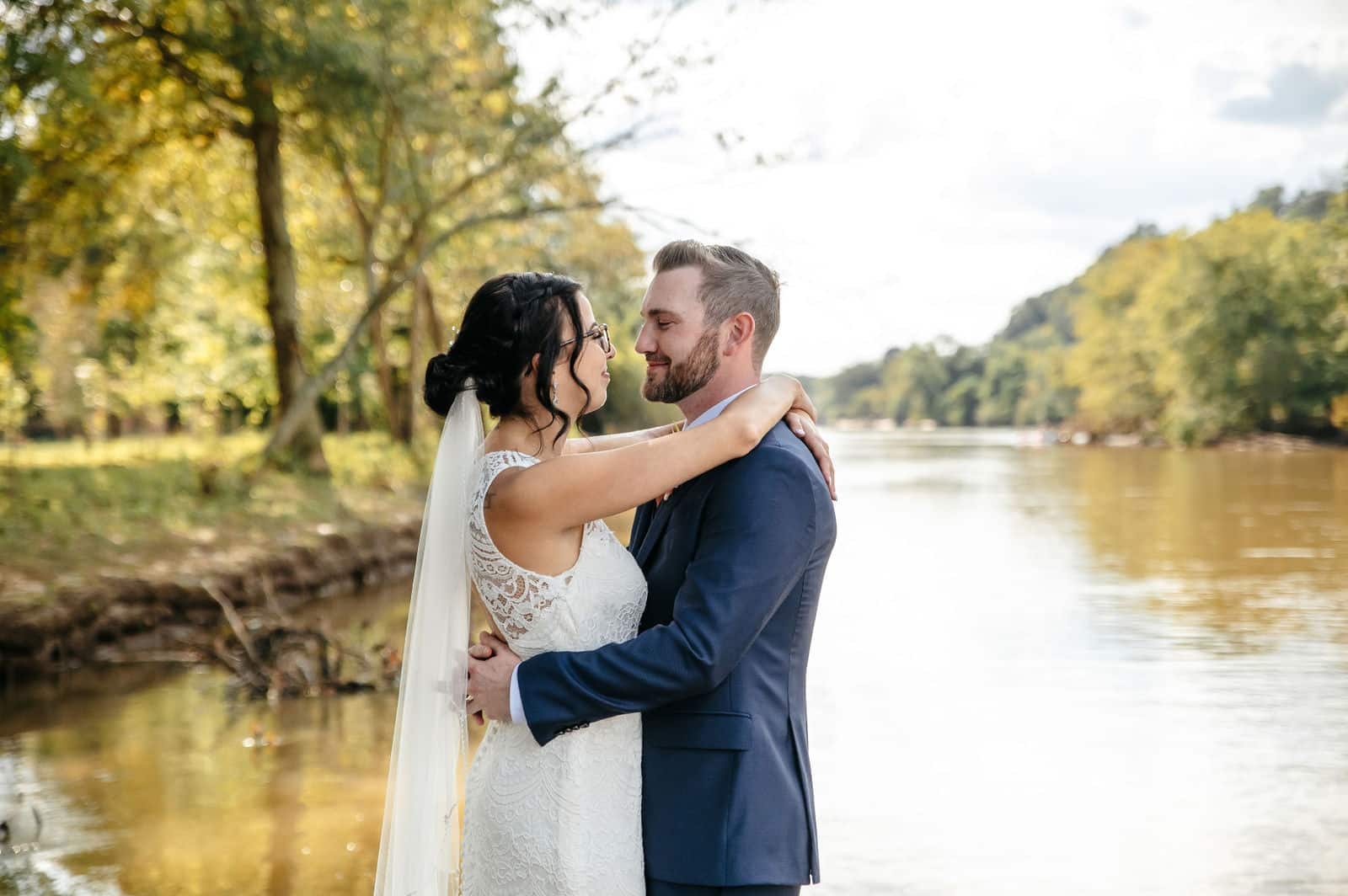 Bride and groom embracing on a riverbank in autumn light.