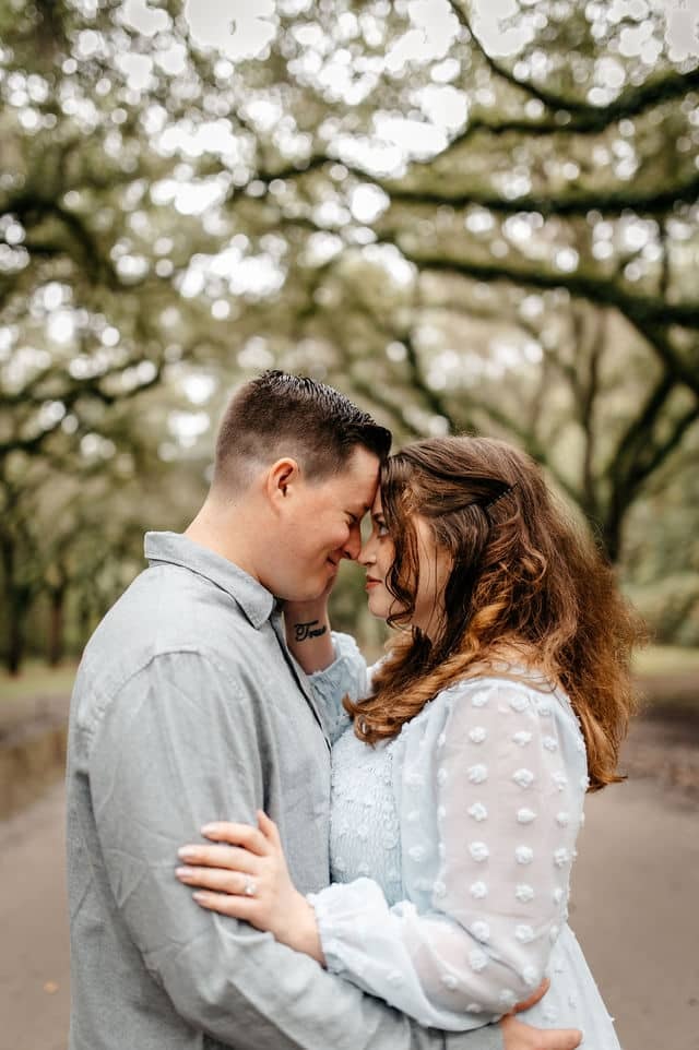 An engaged couple forehead to forehead beneath an avenue of live oaks.