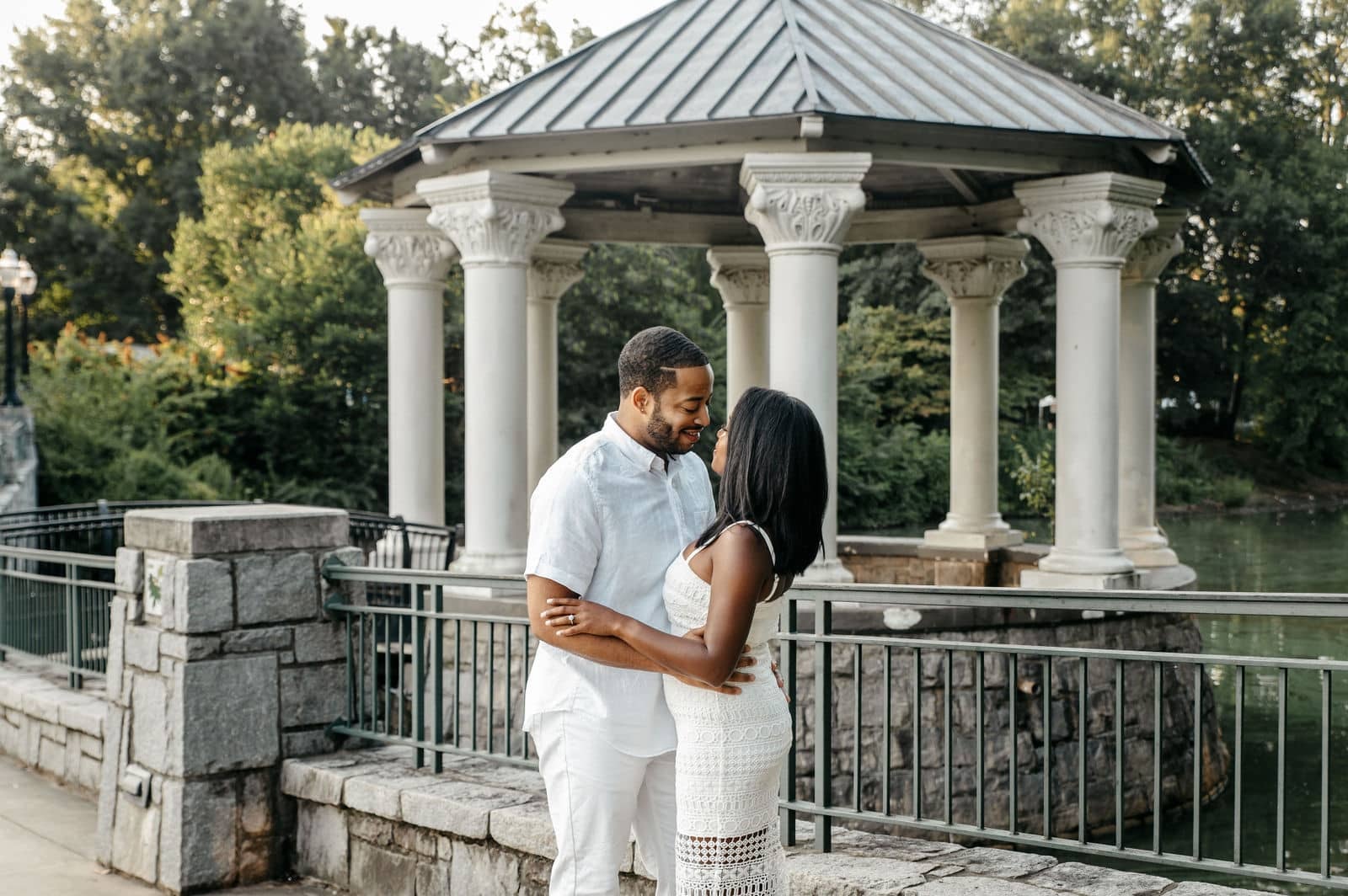 An engaged couple face to face in front of a park gazebo beside the water.