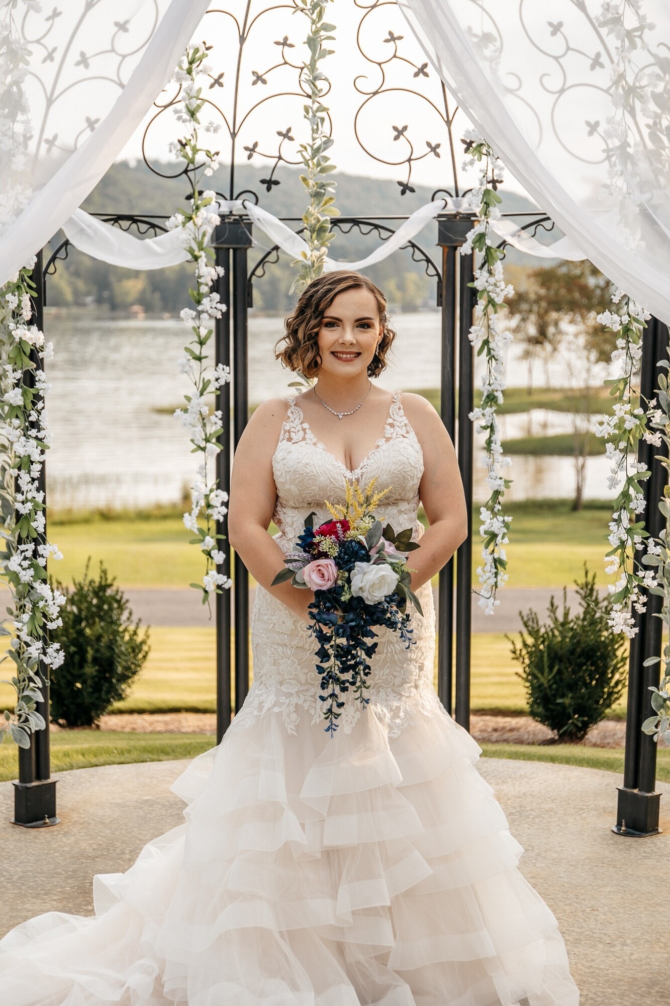 A bride standing alone under a white floral arch beside a lake, holding a bouquet of bright pink and cream flowers.