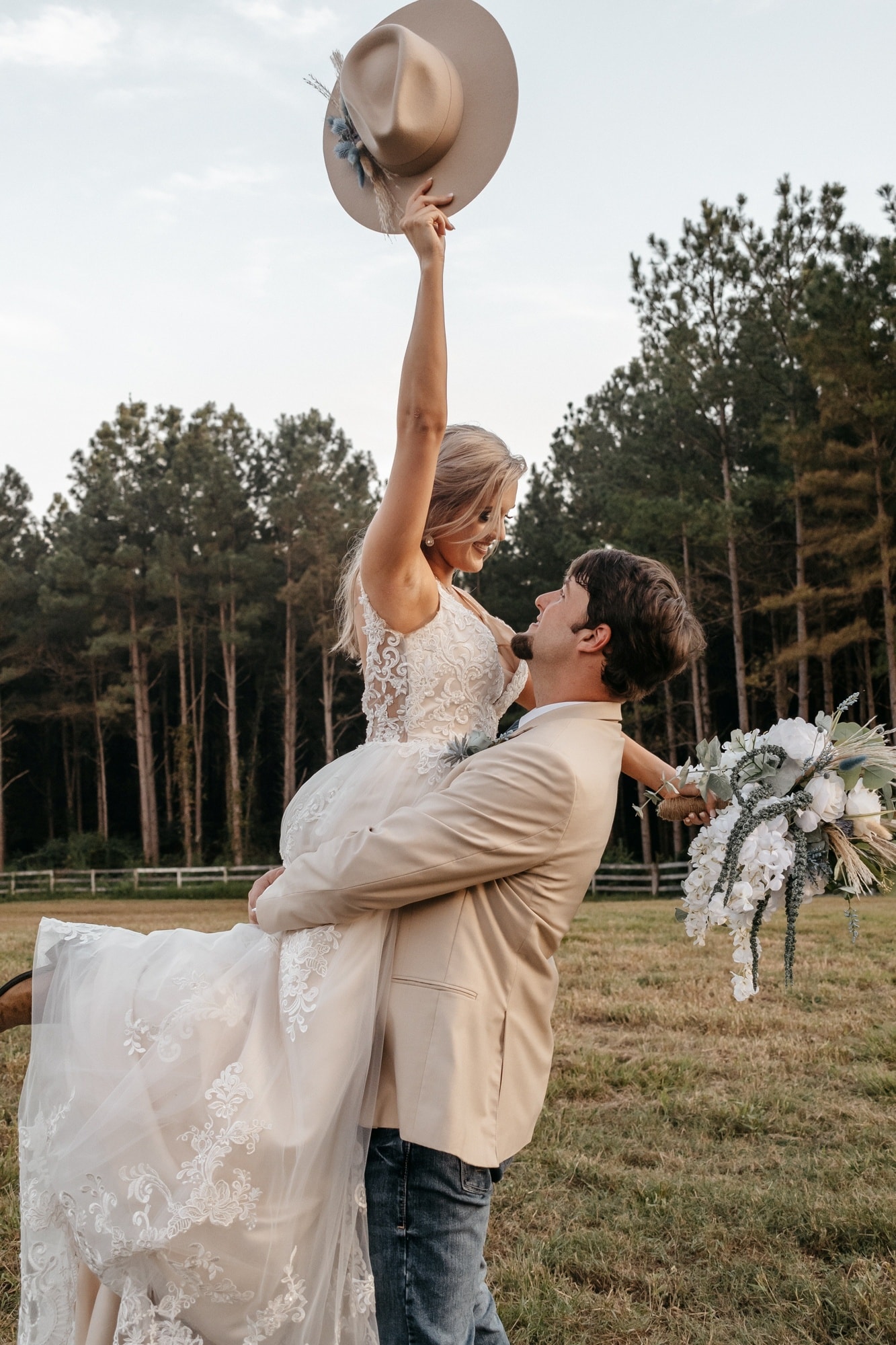 A groom lifting his bride off her feet while she raises his hat overhead, in a field in front of tall pines.