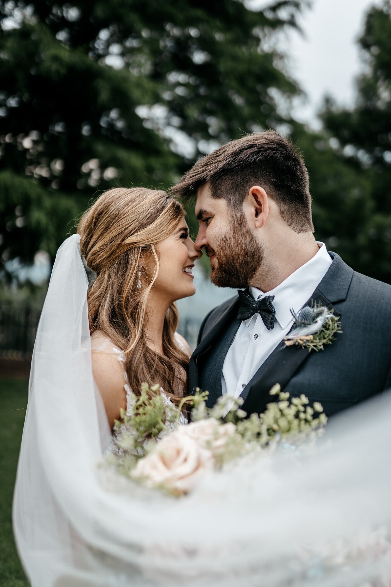 A bride and groom leaning their foreheads together over her bouquet of cream roses and eucalyptus.