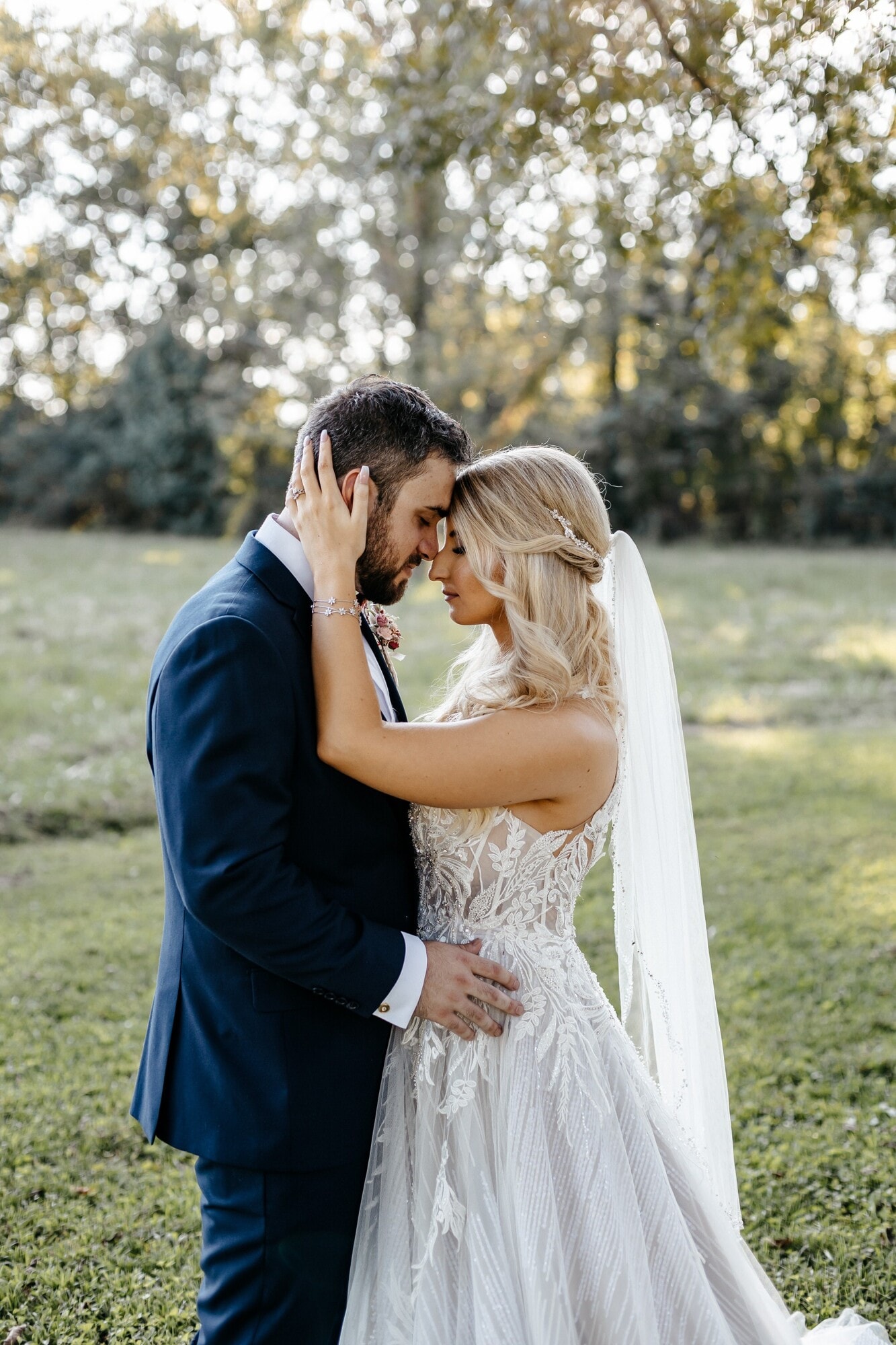 A groom in a navy suit holding his bride's face in both hands, their foreheads together, in a green field at dusk.