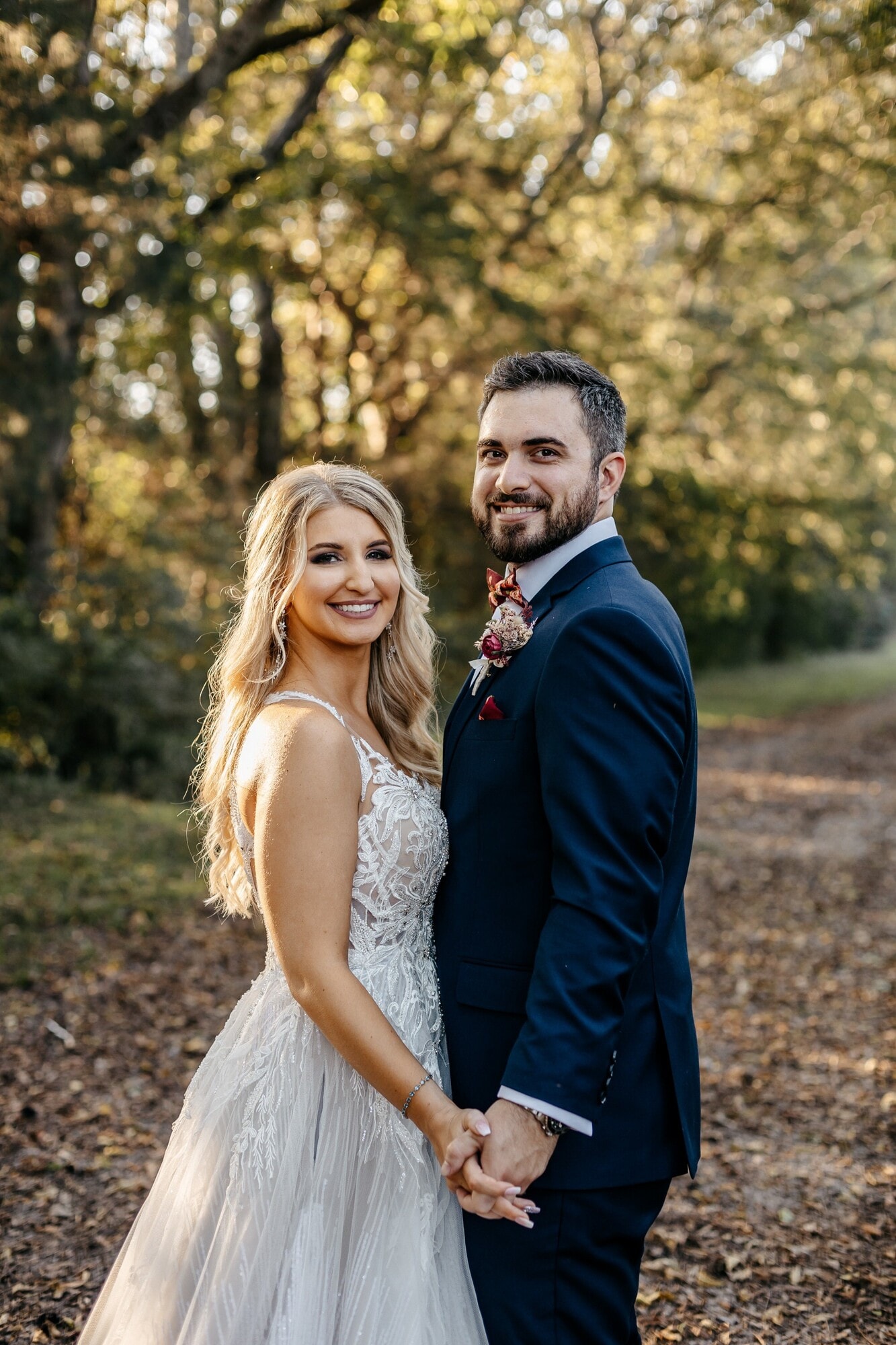 A bride and groom holding hands and smiling at each other on a wooded path in low autumn sunlight.