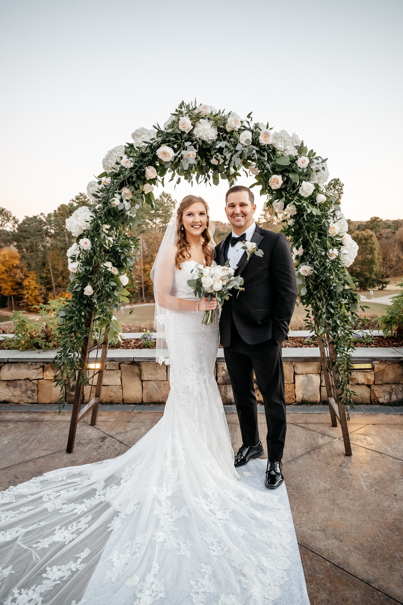 A bride and groom standing together under a white and green floral arch on a stone terrace, her train fanned out across the flagstones.