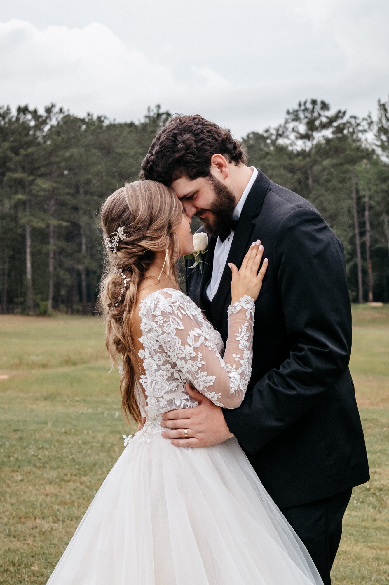 A bride in a long-sleeved lace gown, her hair in a braided updo, resting her hands on her groom's chest on an open lawn.