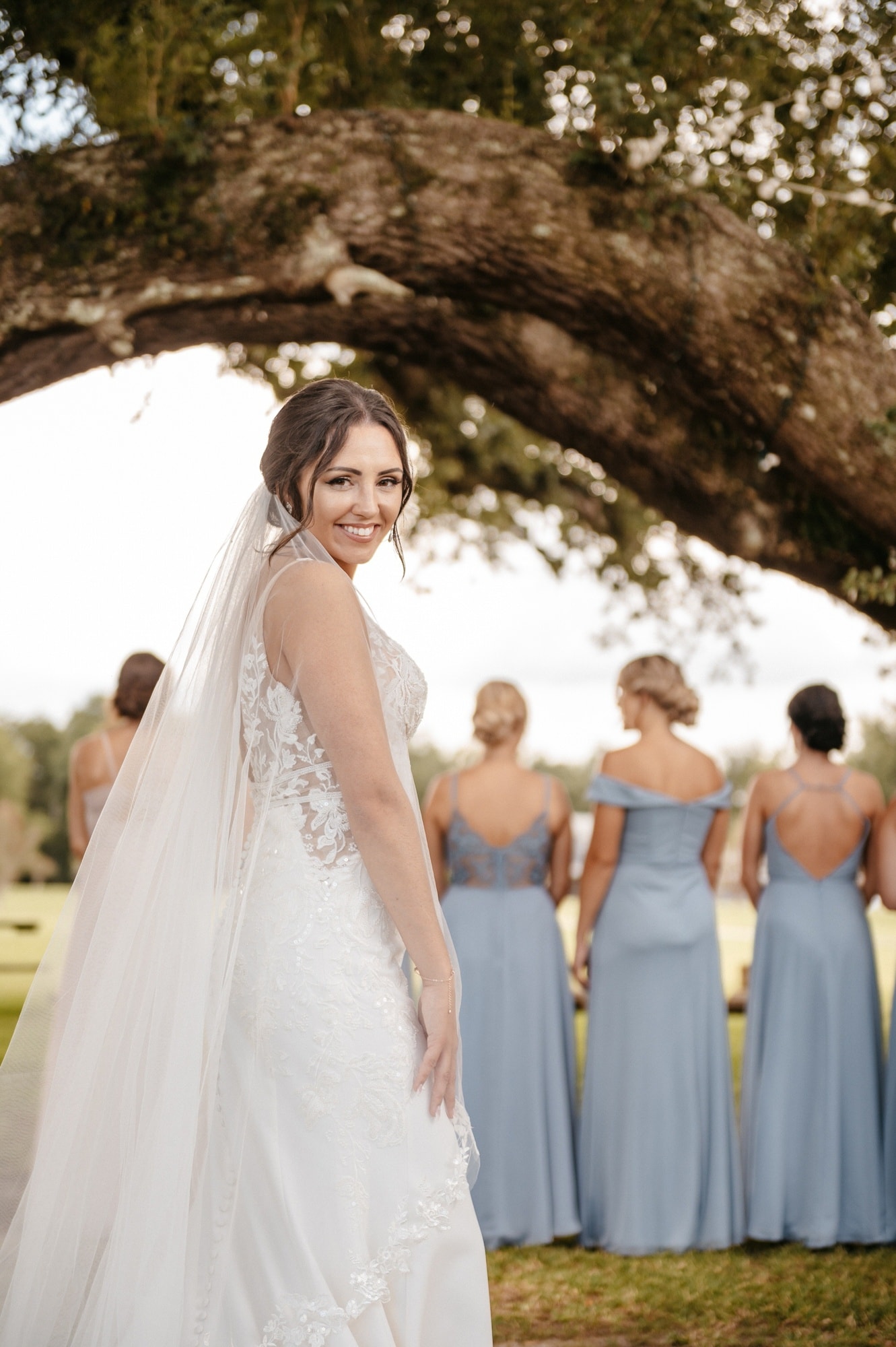 A bride in a long veil glancing back over her shoulder while her bridesmaids, in pale blue, face away under a spreading oak.