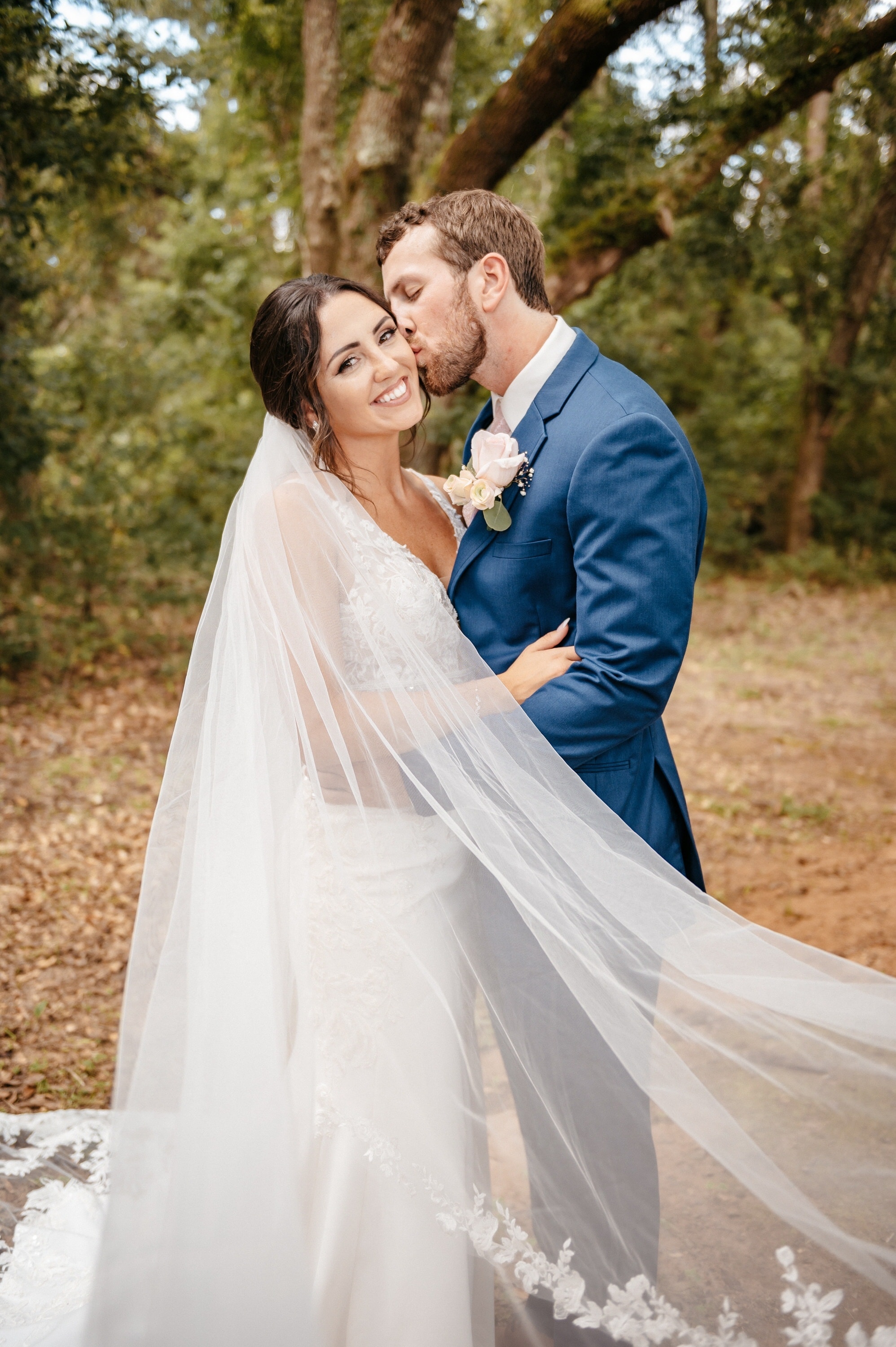 A groom kissing his bride's cheek under trees, her long lace-edged veil sweeping across the frame.