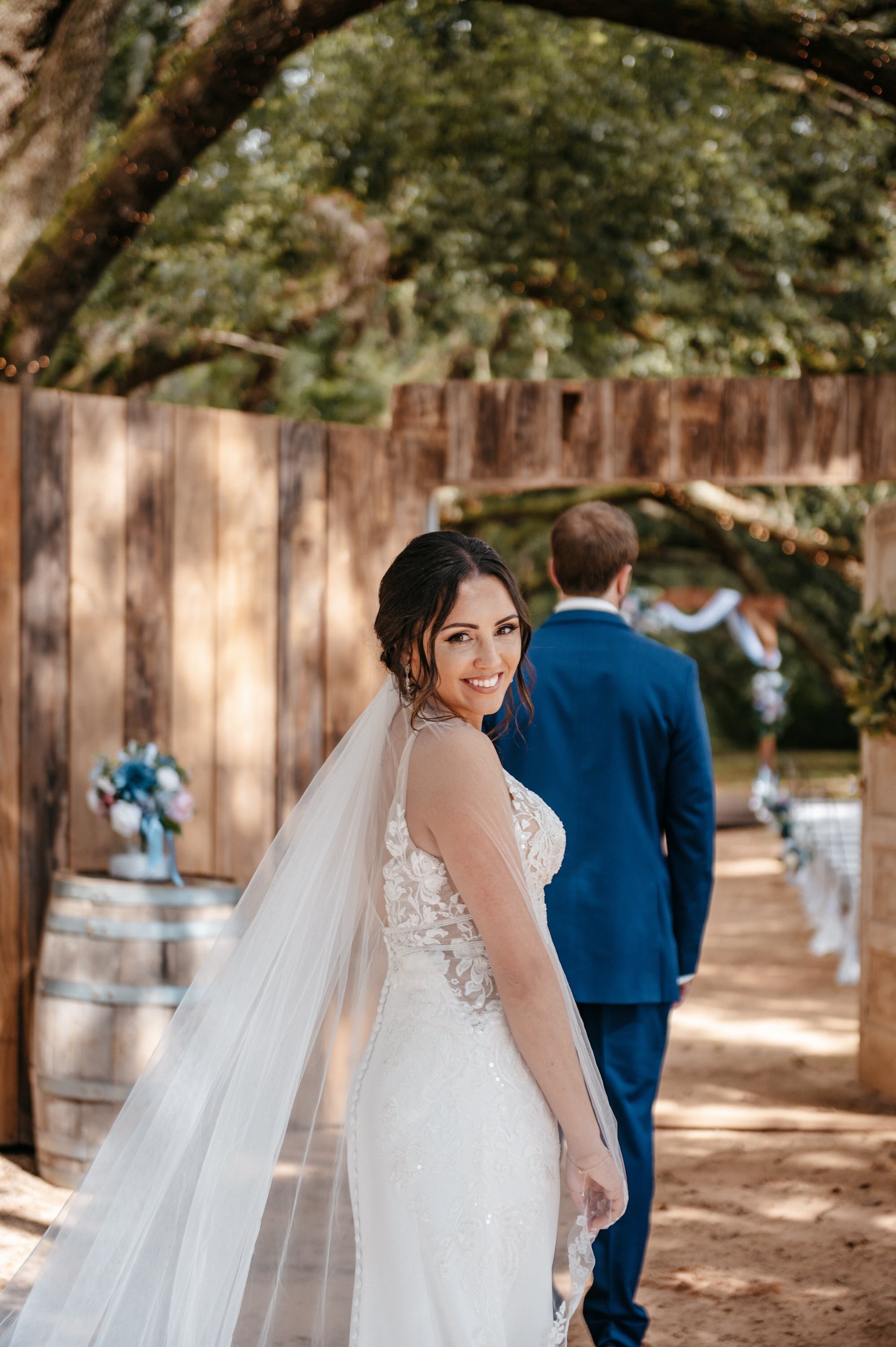 A bride in a cathedral-length veil looking back at the camera while her groom stands facing away beside a wooden fence.