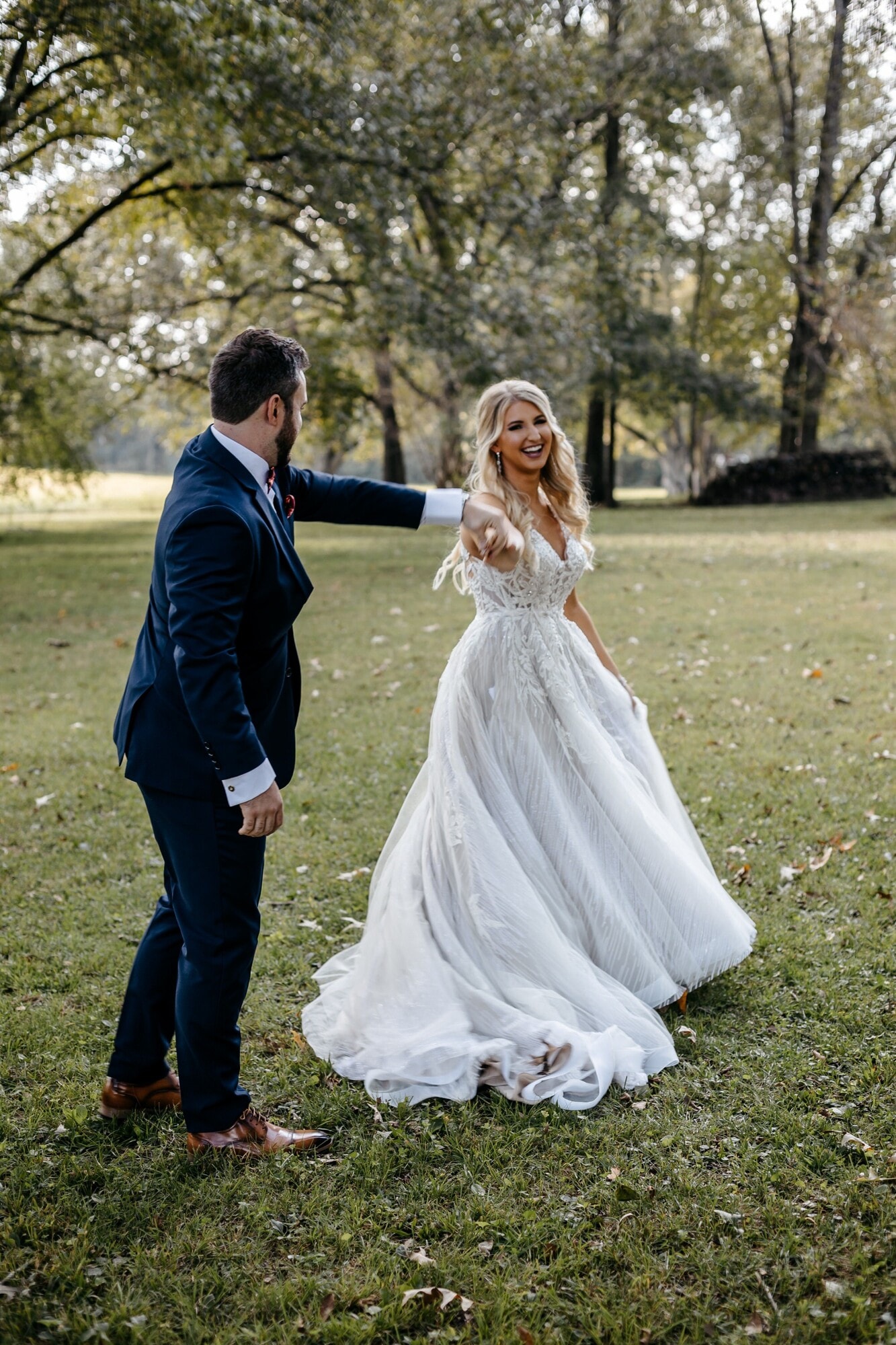 A groom in a navy suit leading his bride by the hand across a lawn, the long skirt of her gown trailing behind her.