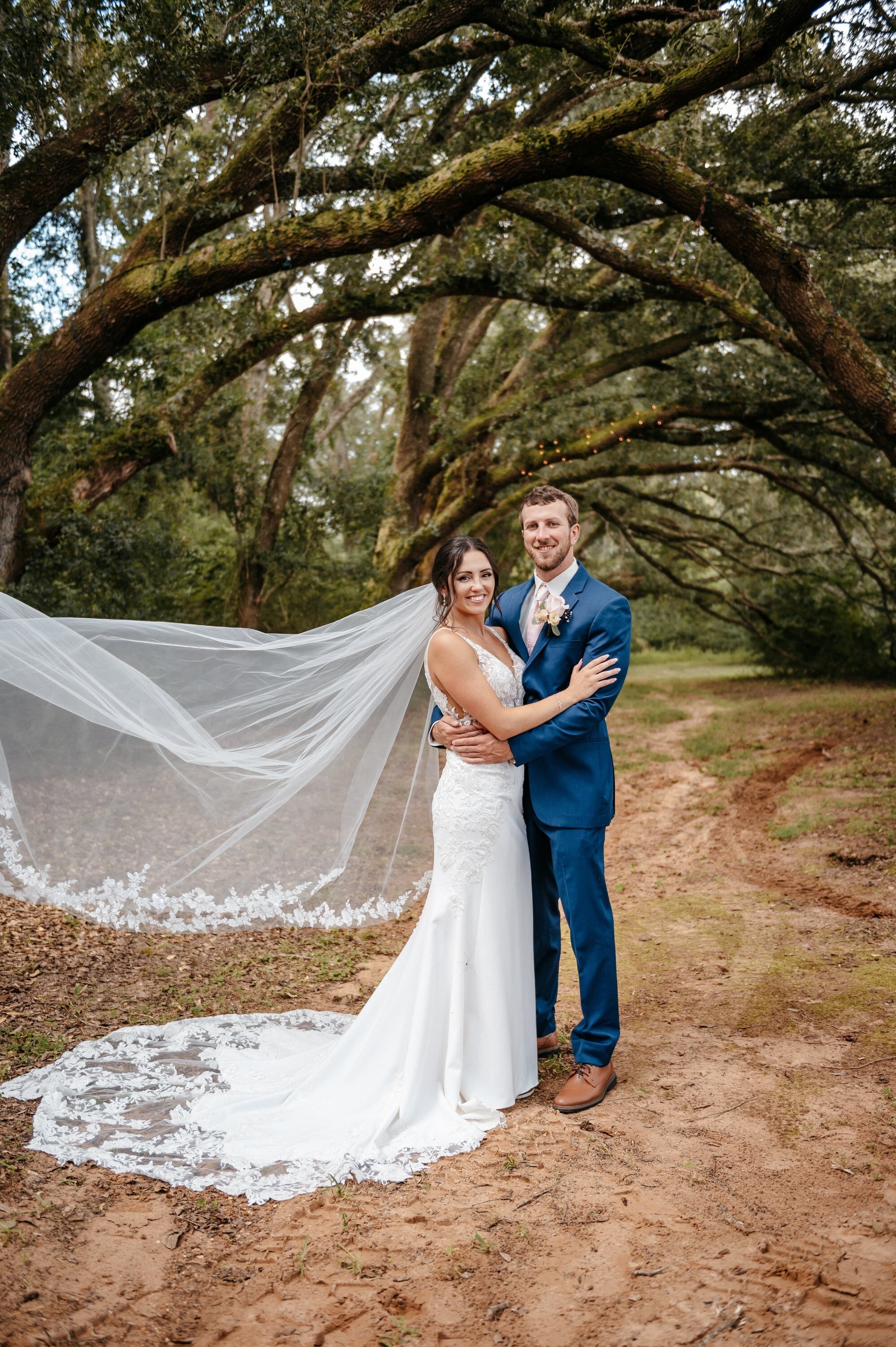 A bride and groom under an avenue of moss-draped oaks, her veil laid out along the sandy path behind her.