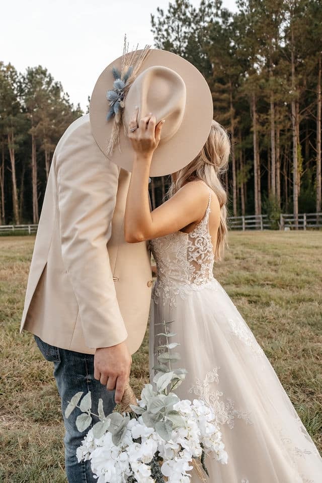 A bride and groom kissing behind a wide-brimmed hat she holds up to hide them, in a field edged with pines.