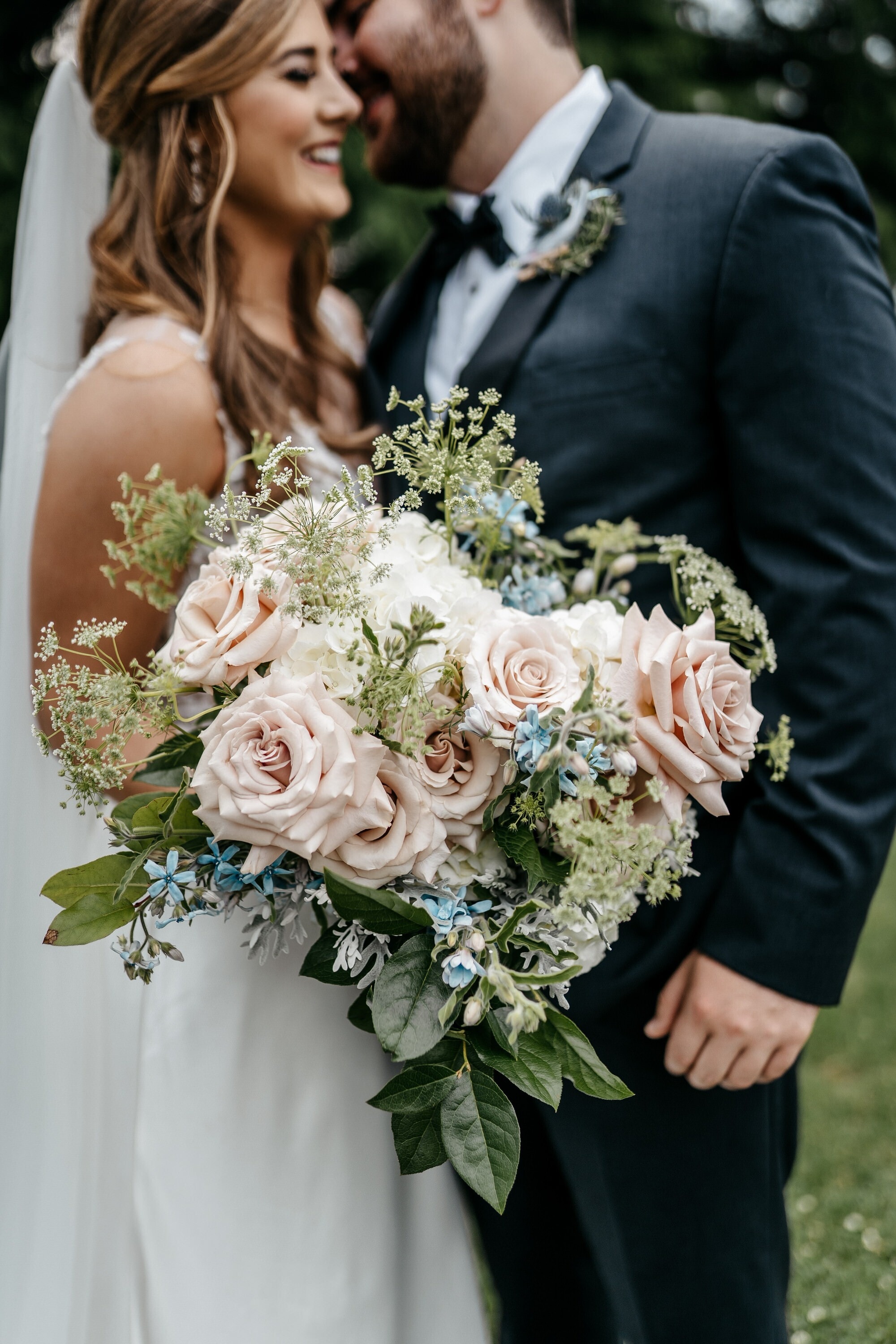A close-up of a bride holding a large bouquet of blush roses and pale blue blooms, her groom beside her in a black tuxedo.