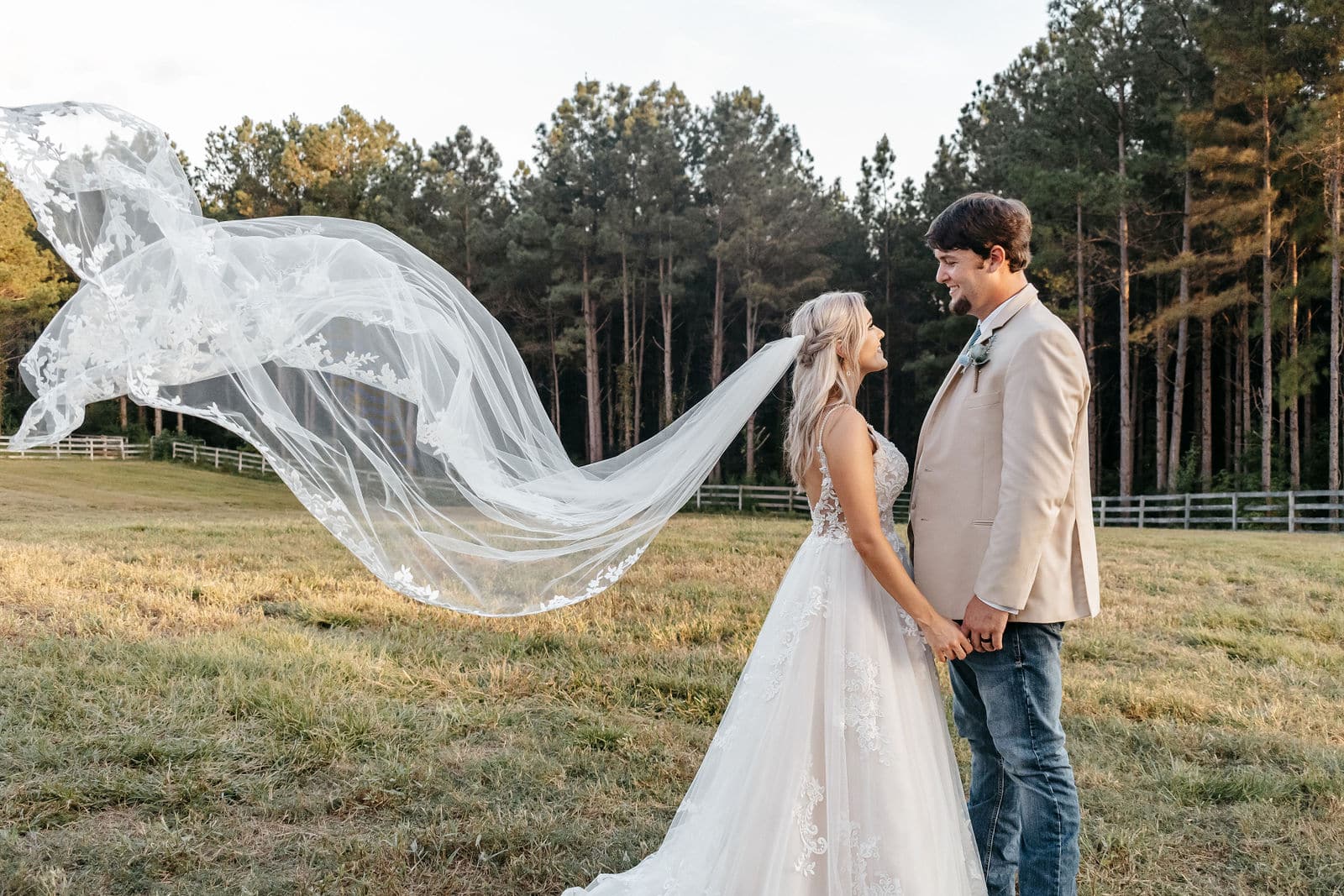 A bride's veil caught by the wind and streaming out behind her as she holds hands with her groom in an open field.