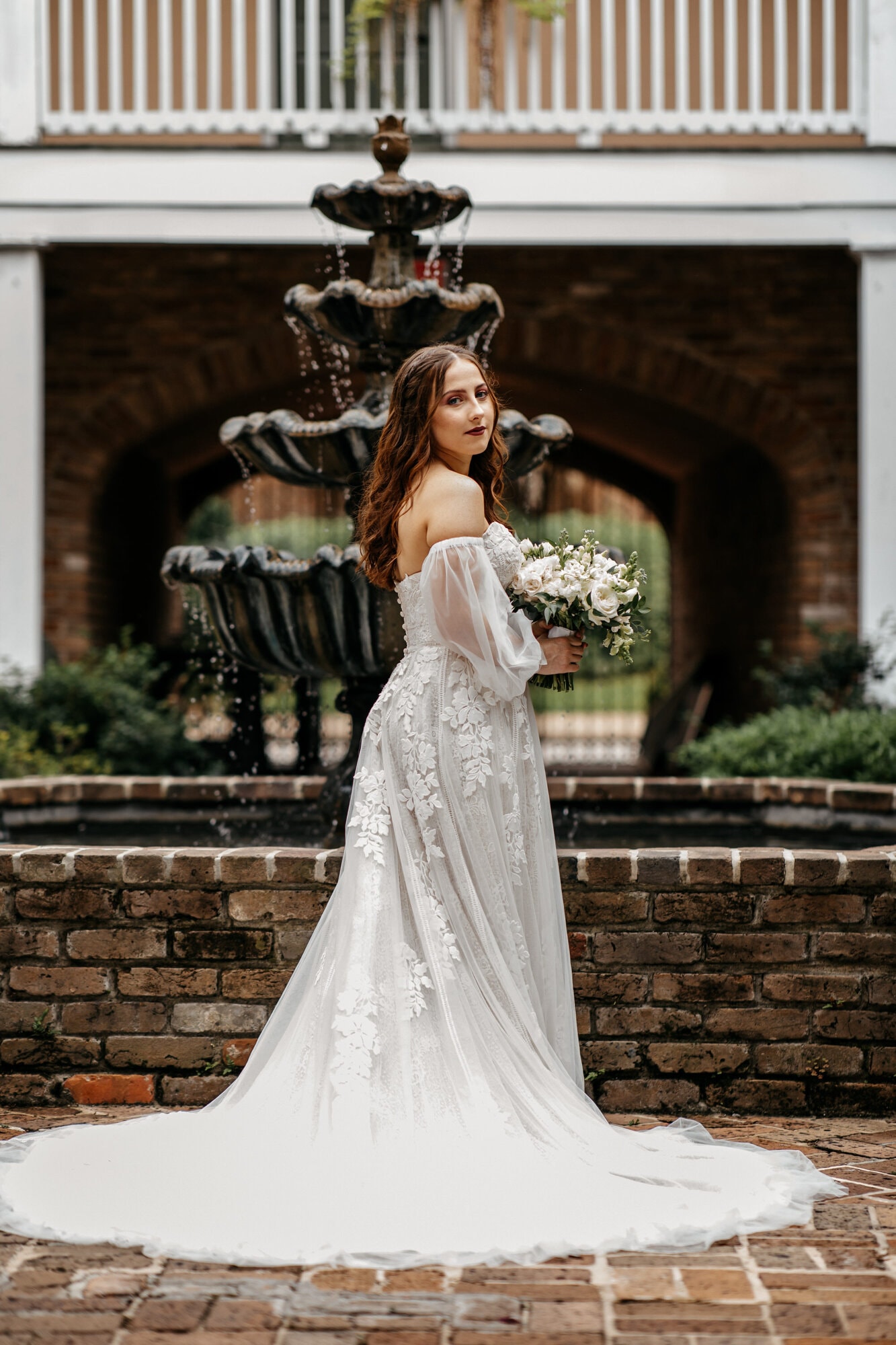 A bride in an off-the-shoulder lace gown standing in front of a brick fountain beneath a covered porch, holding a small bouquet.