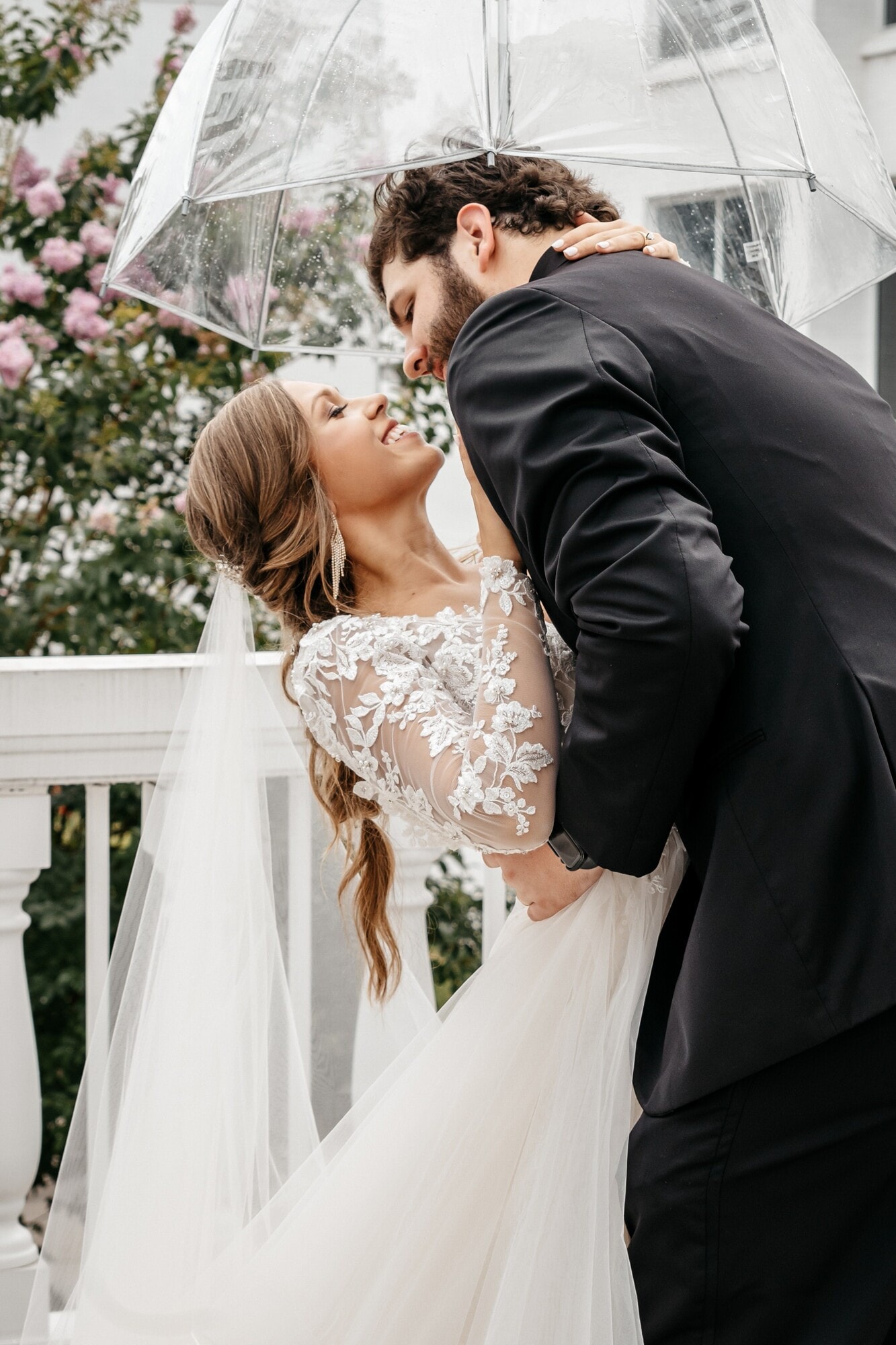 A bride and groom kissing under a clear umbrella beside a white porch rail hung with pink blossom.