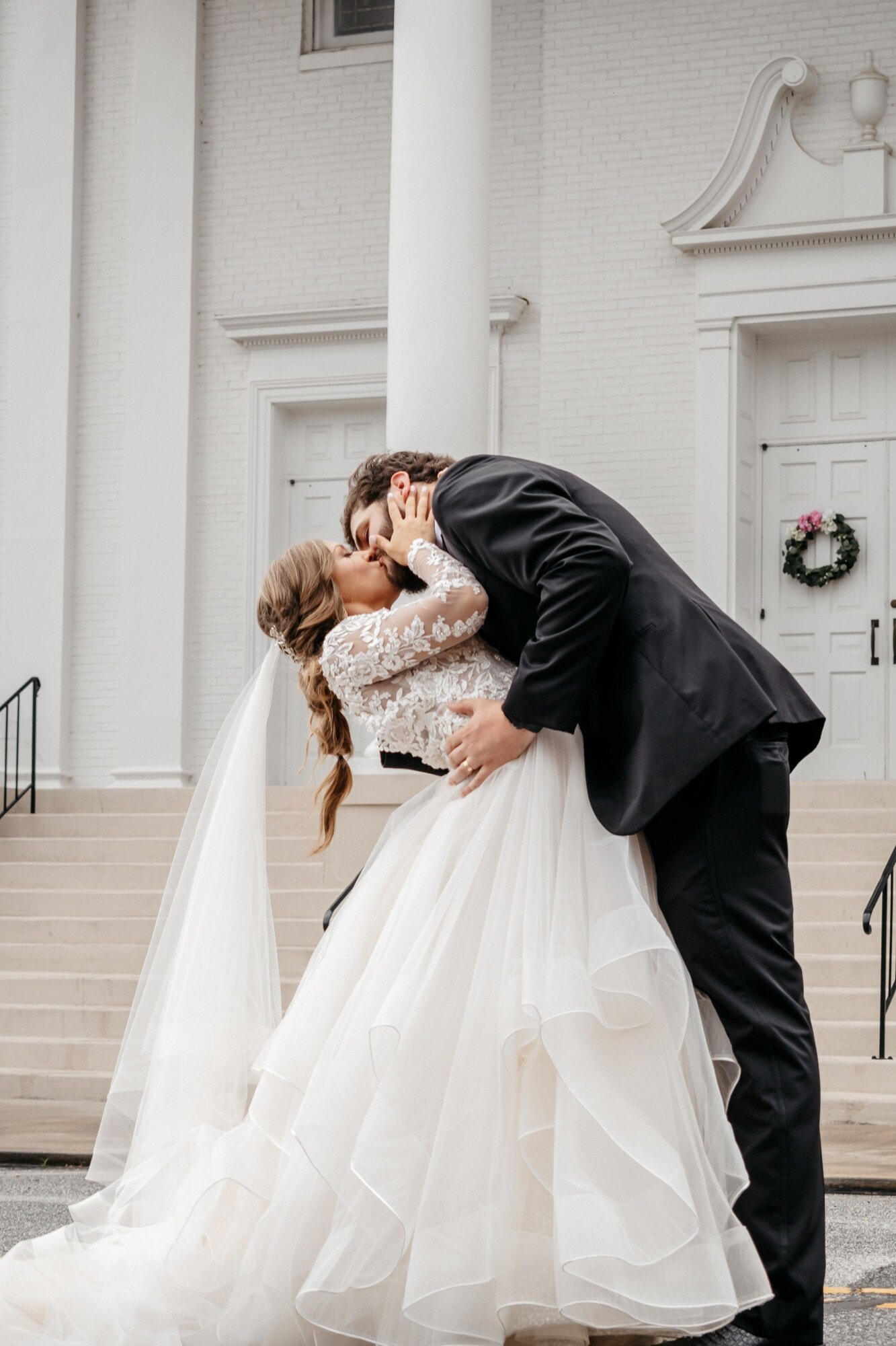 A groom dipping his bride backwards for a kiss on the steps of a white chapel.