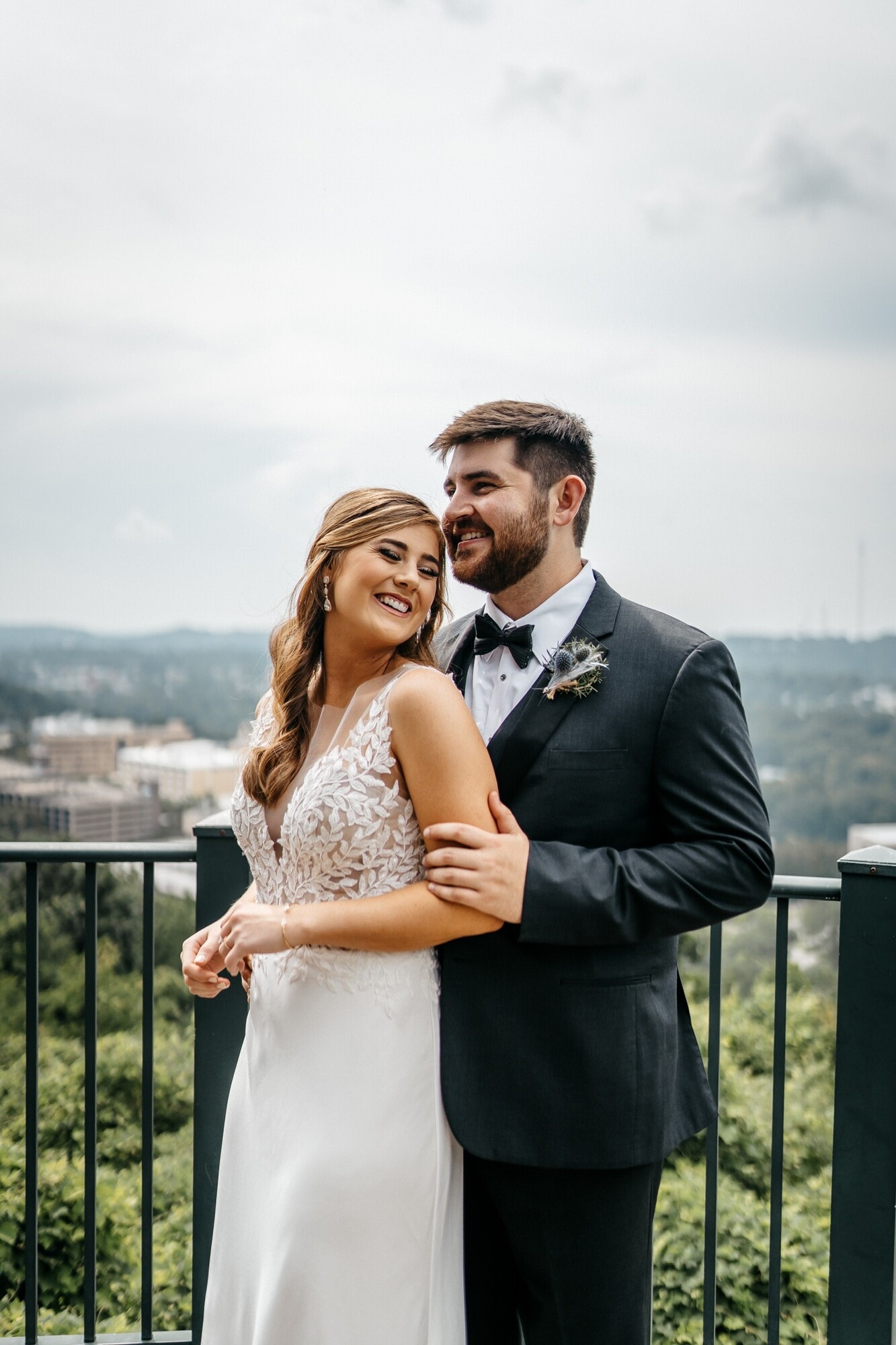 A bride in a lace fit-and-flare gown and a groom in a black tuxedo standing arm in arm at a balcony railing, with a wooded skyline behind them.