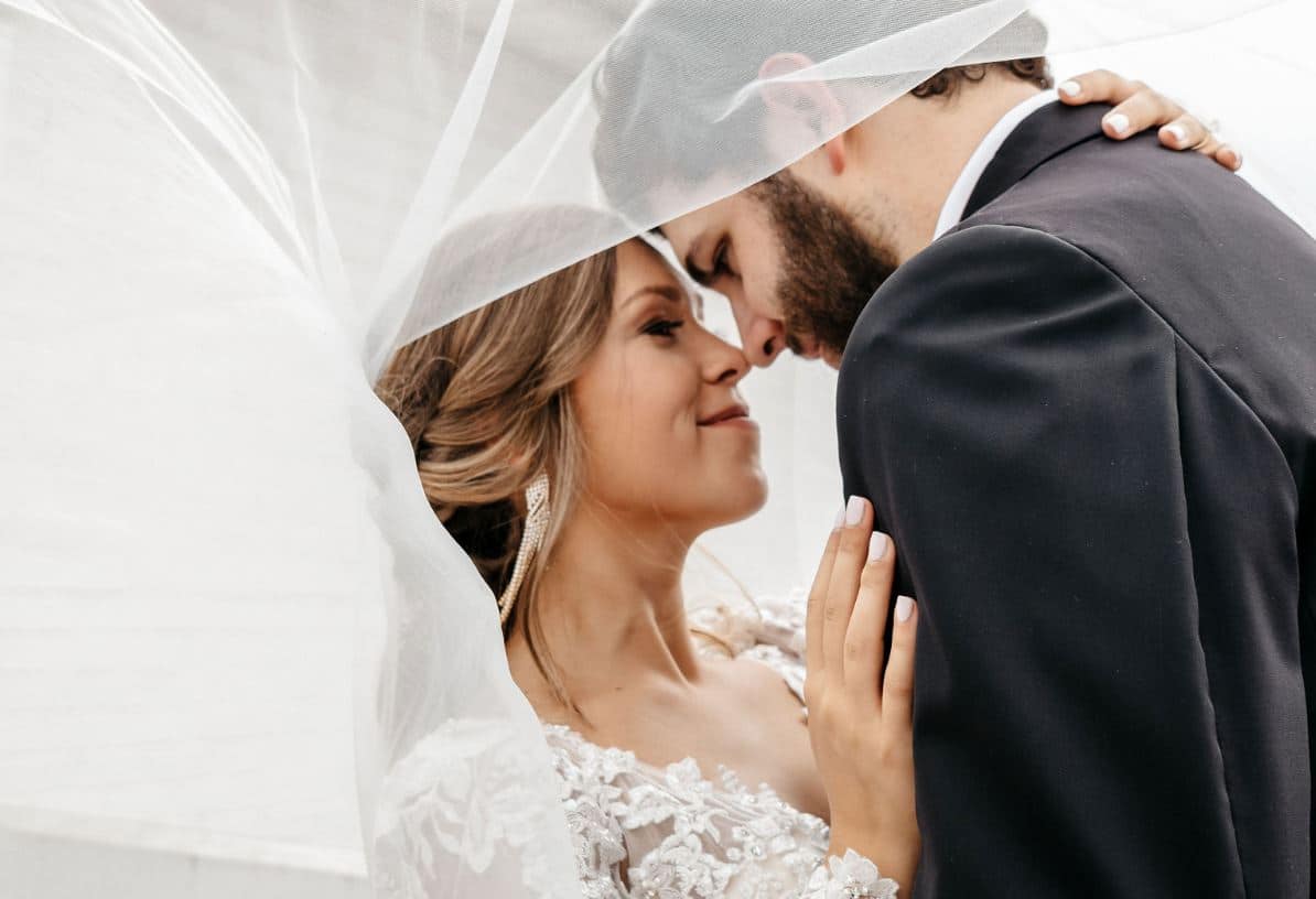 A bride and groom leaning their foreheads together beneath her veil, held up around them both like a canopy.