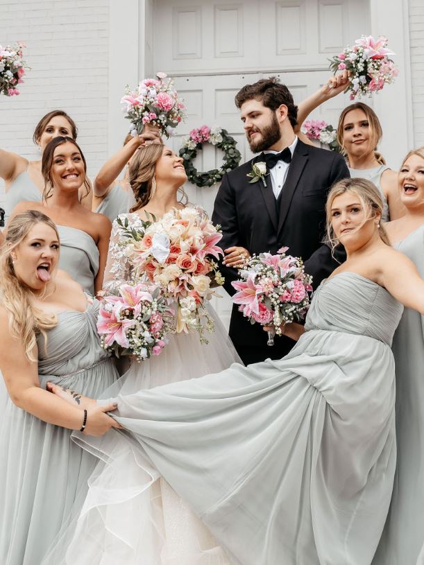 Bridesmaids in sage dresses holding their bouquets up in the air around the laughing bride and groom.