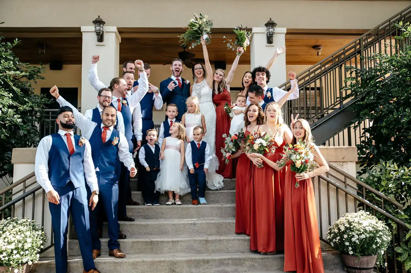 A large wedding party cheering on the steps of a venue, in rust and navy.
