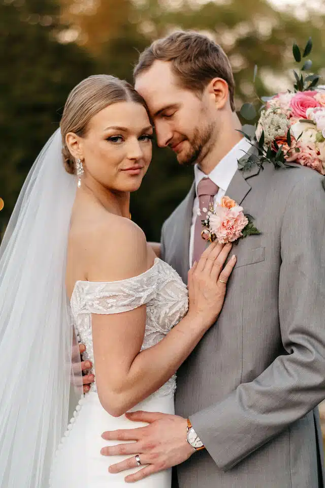 A bride in an off-the-shoulder lace gown and long veil resting her head against her groom, who wears a grey suit with a pink rose boutonniere.