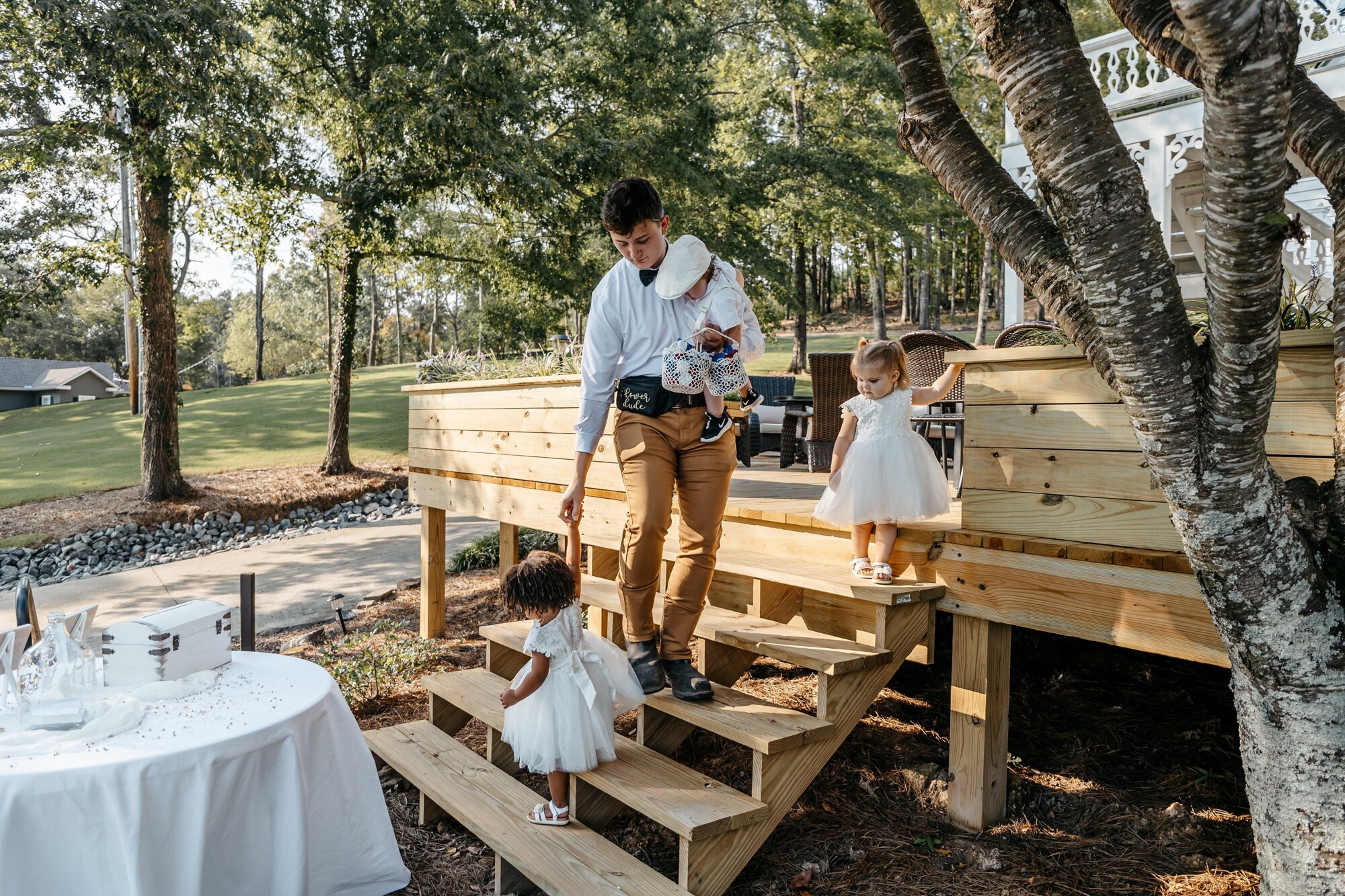 A groomsman holds a baby wearing white and escorts flower girls in white dresses down wooden stairs at a georgia outdoor wedding venue